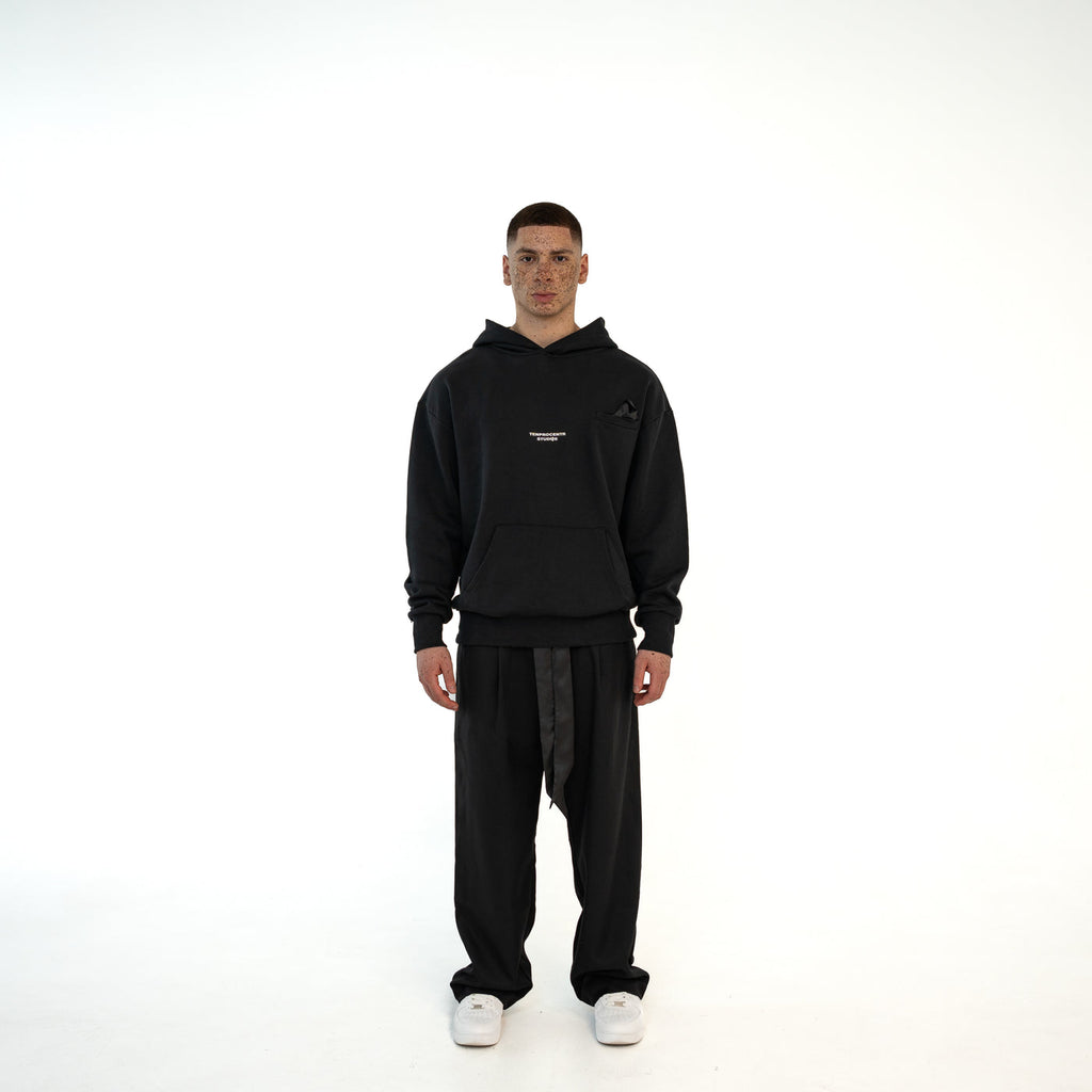 Model traegt TenProcentr BASTILLE Heavyweight Hoodie in Schwarz, 500 GSM, Vorderansicht mit Brustlogo und Kaengurutasche