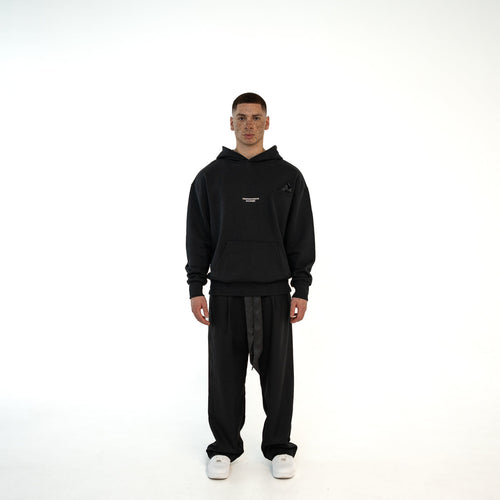 Model traegt TenProcentr BASTILLE Heavyweight Hoodie in Schwarz, 500 GSM, Vorderansicht mit Brustlogo und Kaengurutasche